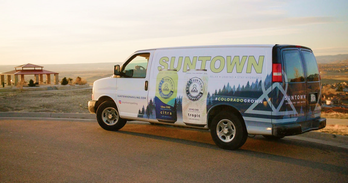 Van wrapped with logo and /assets/wflow of cans in parking lot with overview of mountains behind.